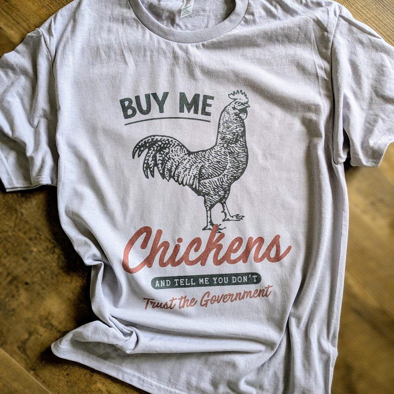 Op de afbeelding: Een lichtgrijs T-shirt met een zwart-witte illustratie van een haan. De tekst "Buy Me Chickens and Tell Me You Don't Trust the Government" staat op het T-shirt gedrukt.