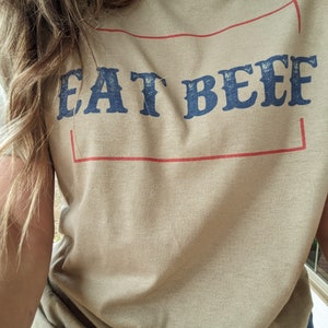May include: A tan t-shirt with a red rectangle around the text "EAT BEEF" in blue lettering.
