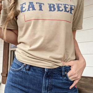 May include: A tan t-shirt with the text "EAT BEEF" in blue lettering inside a red rectangle.