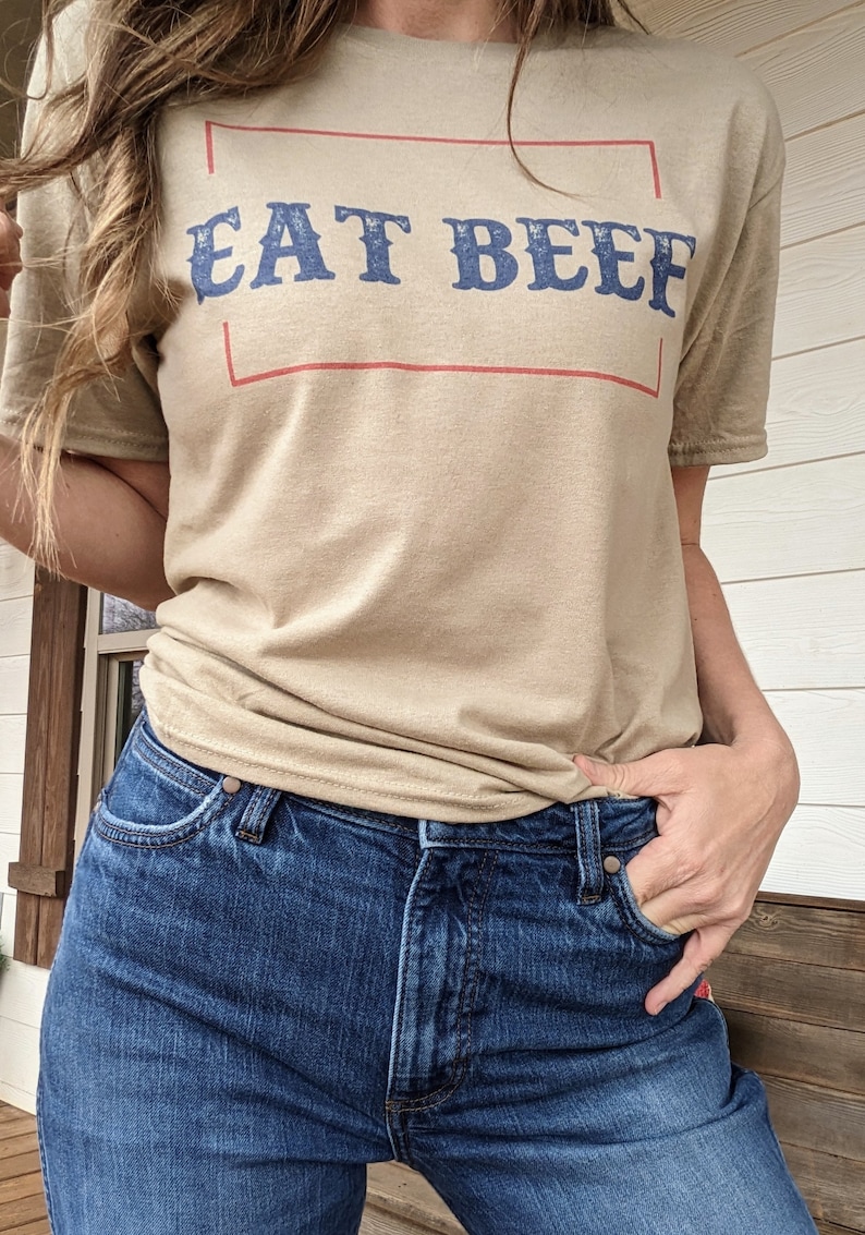 May include: A tan t-shirt with the text "EAT BEEF" in blue lettering inside a red rectangle.