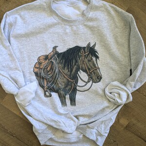 May include: A light gray sweatshirt featuring a detailed illustration of a horse with a saddle and bridle. The horse is depicted in shades of brown and black, with the saddle in a lighter tan color. The sweatshirt has a crew neck and long sleeves.