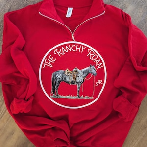 May include: Red quarter-zip sweatshirt with a white circle graphic featuring a horse with a saddle and the text "The Ranchy Roan" in a white circle.