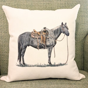 May include: A white cushion featuring a grey horse wearing a brown saddle and a rope halter. The horse is standing on a green field.