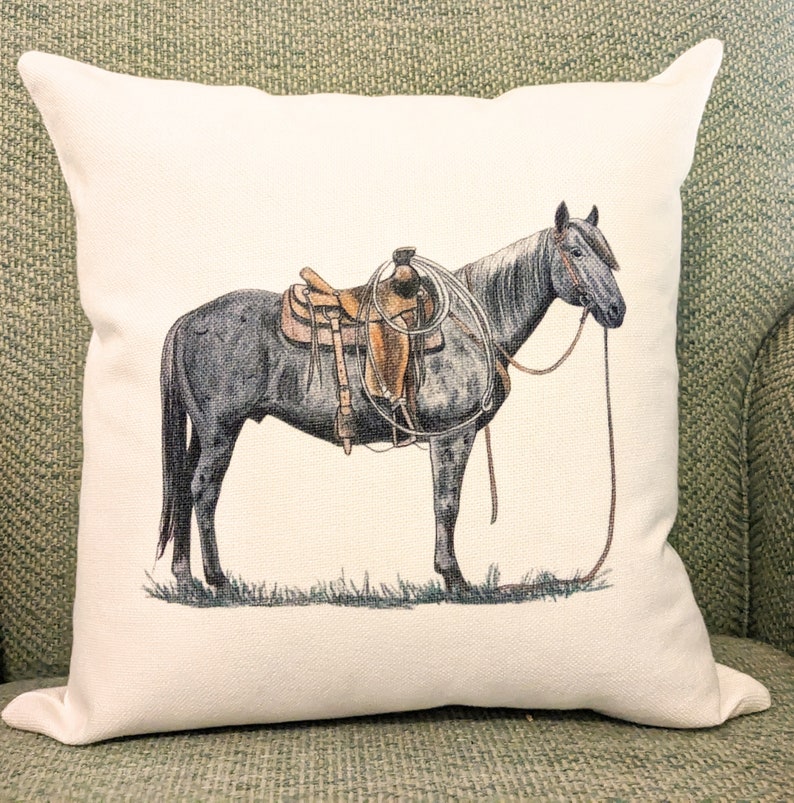 May include: A white cushion featuring a grey horse wearing a brown saddle and a rope halter. The horse is standing on a green field.