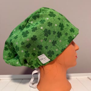 May include: Green scrub hat with a shamrock pattern and silver glitter. The hat has a white tie and a small white label that says "Mimi's Scrubs".