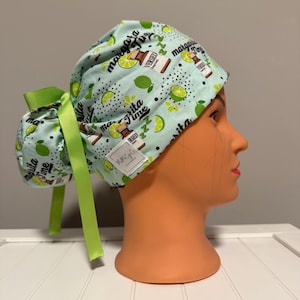 May include: A light blue scrub cap featuring a repeating pattern of lime slices, small medicine vials labeled Versed, and the text Margarita Time with green Z symbols. The cap includes a bright green ribbon tie and a small white rectangular brand tag.