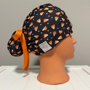 May include: A black scrub cap with a candy corn pattern and white stars. The cap has an orange ribbon tied in a bow. A small white tag with black text is sewn on the side. The cap is displayed on a mannequin head.