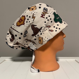 May include: A white scrub hat with a black and white polka dot pattern. The hat features a variety of Harry Potter themed illustrations, including a Hogwarts house crest, a wizard's hat, a lightning bolt, and the word "MAGIC".