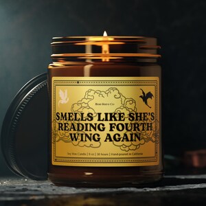 Peut inclure: Un bocal en verre brun contenant une bougie avec une étiquette dorée sur laquelle est inscrit "Smells Like She's Reading Fourth Wing Again". La bougie est allumée et la flamme est visible. L'étiquette comprend également le texte "Bon Horo Co", "Soy Wax Candle | 9 oz | 50 hours | Hand-poured in California".
