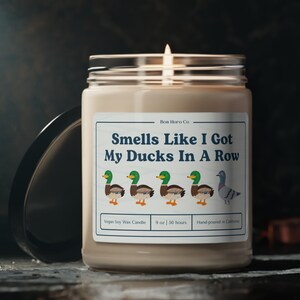 Può includere: Una candela di cera di soia beige in un barattolo di vetro con un'etichetta bianca che recita "Bon Horo Co Smells Like I Got My Ducks In A Row Vegan Soy Wax Candle 9 oz | 50 hours Hand-poured in California". L'etichetta presenta una fila di cinque anatre di cartoni animati e un piccione di cartoni animati.