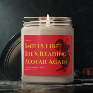 Op de afbeelding: Een brandende, heldere glazen potkaars met een zilveren deksel. De kaars heeft een rood etiket met de gele tekst "SMELLS LIKE SHE'S READING ACOTAR AGAIN". Een zwarte drakenafbeelding staat rechts op het etiket. De kaars is 255 gram.