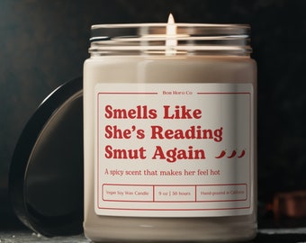 Puede incluir: Una vela beige con una etiqueta roja que dice "Smells Like She's Reading Smut Again" con tres iconos de chiles rojos. La etiqueta también dice "Bon Horo Co", "Vegan Soy Wax Candle", "9 oz | 50 hours", y "Hand-poured in California".