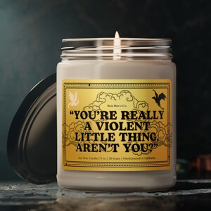 Op de afbeelding: Een brandende sojawas kaars in een heldere glazen pot met een zilveren deksel. De kaars heeft een gouden label met de tekst "YOU'RE REALLY A VIOLENT LITTLE THING, AREN'T YOU?" en decoratieve illustraties. De kaars is 255 g en met de hand gegoten.