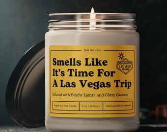 May include: A gold and black candle jar with a label that reads "Smells Like It's Time For A Las Vegas Trip" and "Infused with Bright Lights and Glitzy Casinos". The candle is a vegan soy wax candle and is 9 oz and burns for 50 hours. The candle is hand-poured in California.