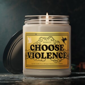 Op de afbeelding: Een brandende sojawas kaars in een heldere glazen pot met een zilveren deksel. Het etiket leest "CHOOSE VIOLENCE" in vette zwarte letters op een gouden achtergrond, met een vleermuis en een gevleugeld wezen. De kaars is 255 gram.