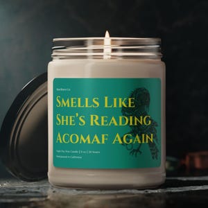 Op de afbeelding: Een brandende, heldere glazen potkaars met een zilveren deksel. De kaars heeft een teal label met de tekst "SMELLS LIKE SHE'S READING ACOMAF AGAIN" in geel. Het label bevat ook een zwarte illustratie van een vogel.
