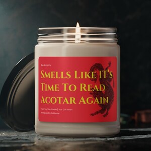 Op de afbeelding: Een brandende sojawas kaars in een heldere glazen pot met een zilveren deksel. Het rode label luidt "Smells Like It's Time To Read ACOTAR Again" met een drakenillustratie. De kaars is 255 gram.