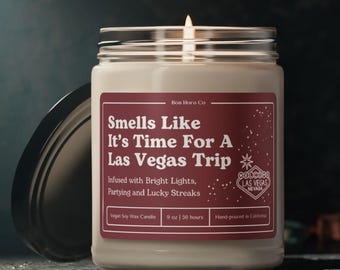 May include: A red and white candle jar with the text "Smells Like It's Time For A Las Vegas Trip" and "Infused with Bright Lights, Partying and Lucky Streaks". The candle is 9 ounces and has a burn time of 50 hours. The candle is hand-poured in California.