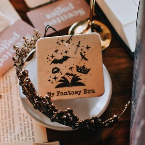 May include: A white ceramic car air freshener with a black and white illustration of a book, a crown, a bat, a dagger, and stars. The text "My Fantasy Era" is printed on the bottom of the air freshener.