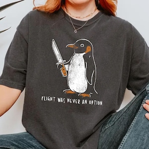 May include: A charcoal gray short-sleeve t-shirt featuring a graphic of a penguin holding a knife with the text "FLIGHT WAS NEVER AN OPTION" printed below. The shirt has a crew neckline and a relaxed fit, with the color name "PEPPER" visible.
