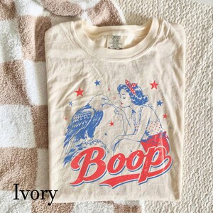 May include: An ivory-colored t-shirt featuring a graphic print of a woman with a red polka-dot hair bow touching the beak of a bald eagle. The design includes red and blue stars and the word Boop in large red letters with a blue outline.