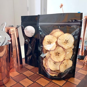 Op de afbeelding: Een close-up van een zwarte hersluitbare zak gevuld met gedroogde appelschijfjes. De zak ligt op een houten snijplank naast een koperen cocktailshaker, een koperen jigger en een koperen cocktailmuddle.