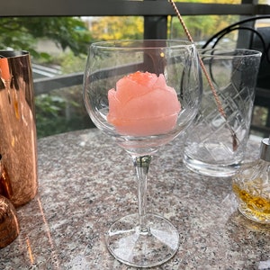 May include: A clear glass with a pink rose-shaped ice cube inside. The glass is sitting on a granite worktop.
