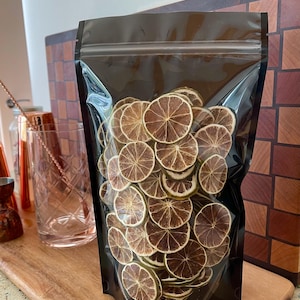 May include: A black bag with a clear window, packed with dried lime slices. The slices are circular, light brown with darker brown centres. The bag rests on a wooden chopping board. Cocktail shaker and bar tools are visible in the background.
