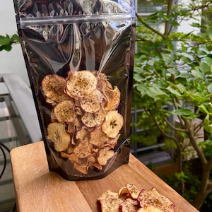 Op de afbeelding: Een doorzichtige plastic zak gevuld met gedroogde appelchips. De chips zijn lichtbruin van kleur met wat rood en zijn bestrooid met kaneel. De zak ligt op een houten oppervlak.