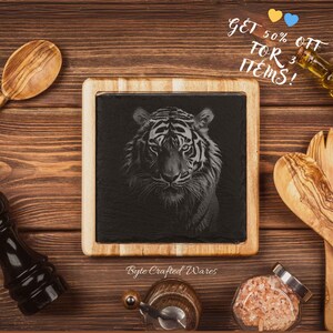 Op de afbeelding: Een zwart leisteen serveerbord met een houten frame heeft een witte tijger gegraveerd in het oppervlak. Het bord is omringd door keukengerei, waaronder een houten lepel, een pepermolen, een zoutvaatje en een fles olijfolie. De tekst "Get 50% off for 3 items!" is in wit op een kleurrijke achtergrond geschreven.