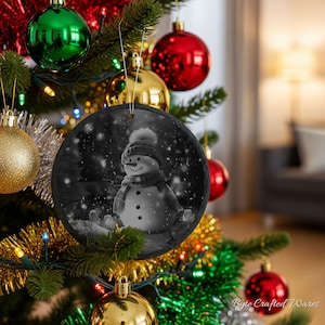May include: A round, black and white slate ornament featuring a snowman wearing a hat and scarf, depicted in a snowy scene. The ornament is suspended by a silver string and hangs from a green Christmas tree decorated with various colorful baubles.