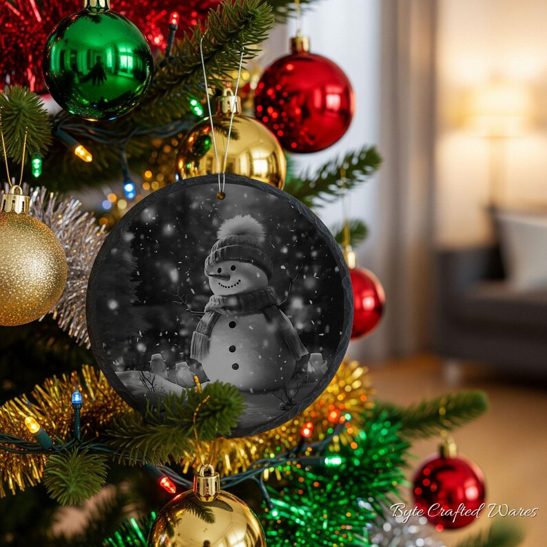 May include: A round, black and white slate ornament featuring a snowman wearing a hat and scarf, depicted in a snowy scene. The ornament is suspended by a silver string and hangs from a green Christmas tree decorated with various colorful baubles.
