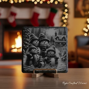 May include: A square slate plaque featuring a black and white illustration of a family of five snowmen standing in a snowy landscape with a house in the background. The snowmen wear hats and scarves. The plaque rests on a gold metal stand.