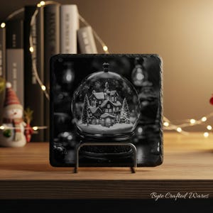 May include: A square black slate coaster featuring a printed graphic of a snow globe containing a winter village scene with houses and pine trees. The coaster is held upright by a silver metal stand, displaying a detailed monochrome winter landscape.