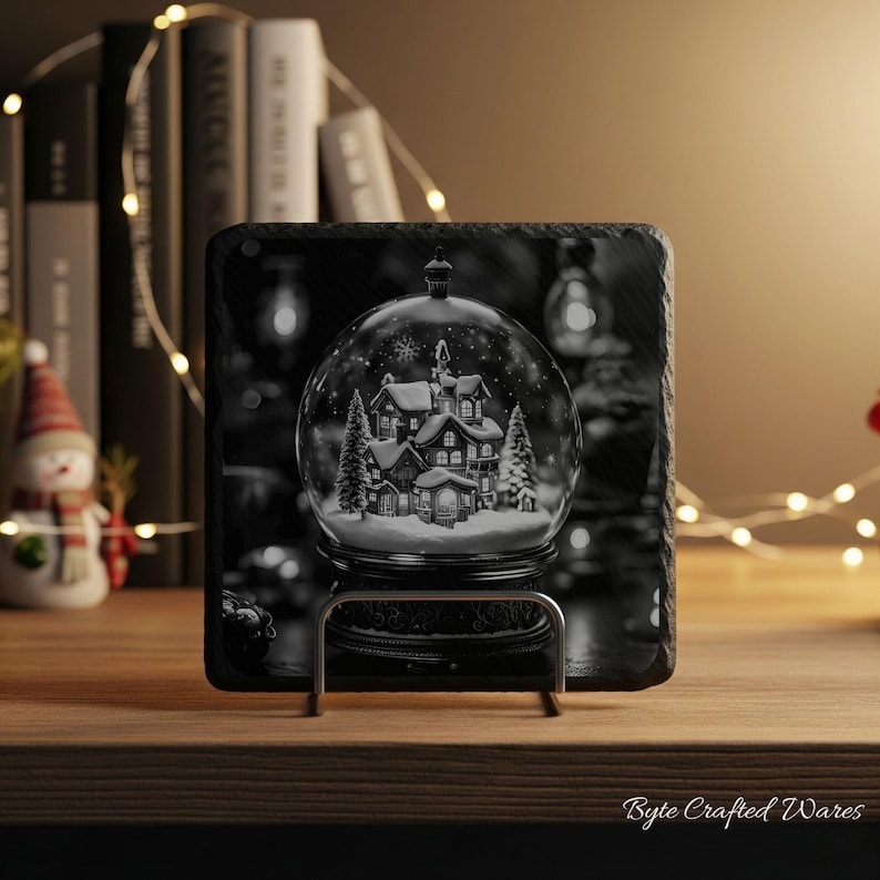 May include: A square black slate coaster featuring a printed graphic of a snow globe containing a winter village scene with houses and pine trees. The coaster is held upright by a silver metal stand, displaying a detailed monochrome winter landscape.