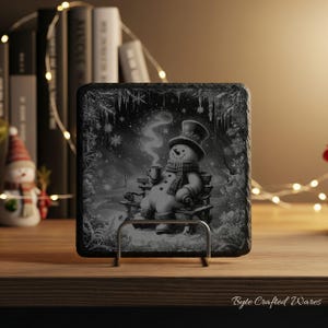 May include: A square black slate coaster featuring a detailed monochrome illustration of a snowman wearing a top hat and scarf, sitting on a bench while holding a steaming mug. The scene is framed by intricate snowflake and icicle patterns.