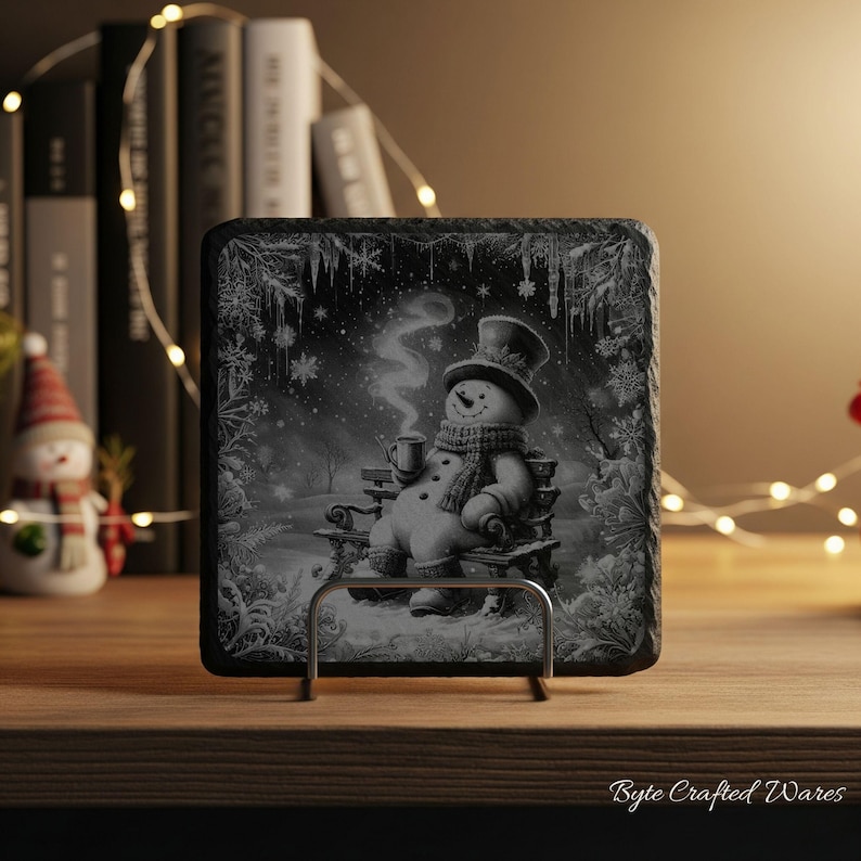 May include: A square black slate coaster featuring a detailed monochrome illustration of a snowman wearing a top hat and scarf, sitting on a bench while holding a steaming mug. The scene is framed by intricate snowflake and icicle patterns.