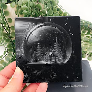 May include: A square black slate coaster featuring a detailed monochrome illustration of a snow globe containing a house and pine trees. The scene inside the globe shows a winter landscape with falling snow. The text Byte Crafted Wares is visible.