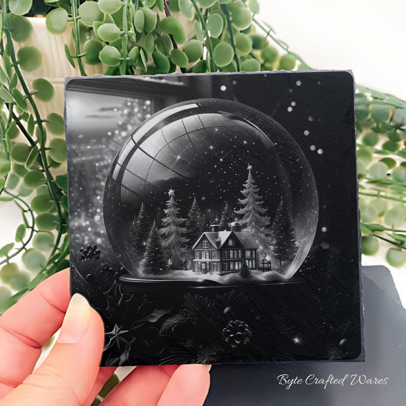 May include: A square black slate coaster featuring a detailed monochrome illustration of a snow globe containing a house and pine trees. The scene inside the globe shows a winter landscape with falling snow. The text Byte Crafted Wares is visible.