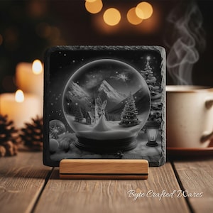 May include: A square slate coaster featuring a black and white illustration of a snow globe containing a church, a fairy, and pine trees against a mountain backdrop. The coaster rests on a light-colored wooden stand with a textured, rough edge.
