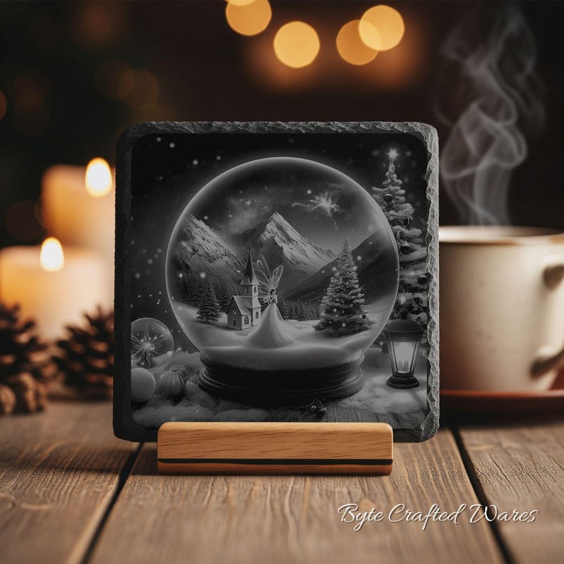 May include: A square slate coaster featuring a black and white illustration of a snow globe containing a church, a fairy, and pine trees against a mountain backdrop. The coaster rests on a light-colored wooden stand with a textured, rough edge.