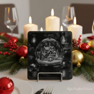 May include: A square black slate coaster featuring a monochromatic illustration of a snow globe containing a winter village scene with houses and pine trees, displayed on a silver metal stand. The text Byte Crafted Wares is visible.