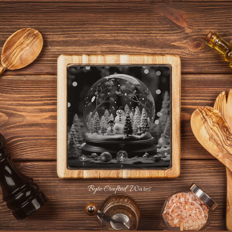 May include: A square wooden trivet featuring a black and white graphic of a snow globe containing two snowmen and pine trees. The text Byte Crafted Wares is printed in a cursive font below the central image on the wooden frame surface.