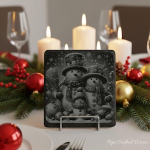 May include: A square slate coaster featuring a black and white illustration of a family of three snowmen wearing hats and scarves, surrounded by snowflakes, ornaments, and pine branches, displayed on a metal stand on a table.