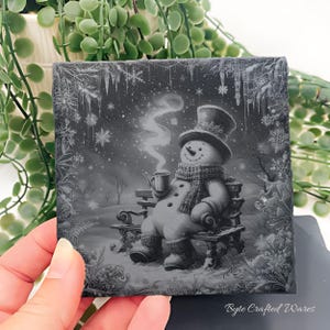 May include: A square slate coaster featuring a grayscale illustration of a snowman wearing a top hat, scarf, and boots, sitting on a bench while holding a steaming mug. The border is decorated with intricate snowflake and icicle patterns.