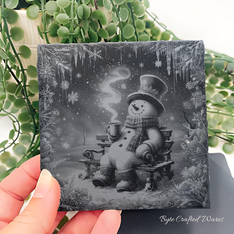 May include: A square slate coaster featuring a grayscale illustration of a snowman wearing a top hat, scarf, and boots, sitting on a bench while holding a steaming mug. The border is decorated with intricate snowflake and icicle patterns.