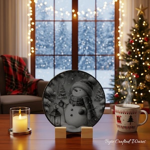 May include: A circular decorative plate featuring a grayscale illustration of a snowman wearing a hat and scarf, standing next to a lantern in a snowy forest. The plate is displayed on a wooden stand between a glass candle holder and a white mug.