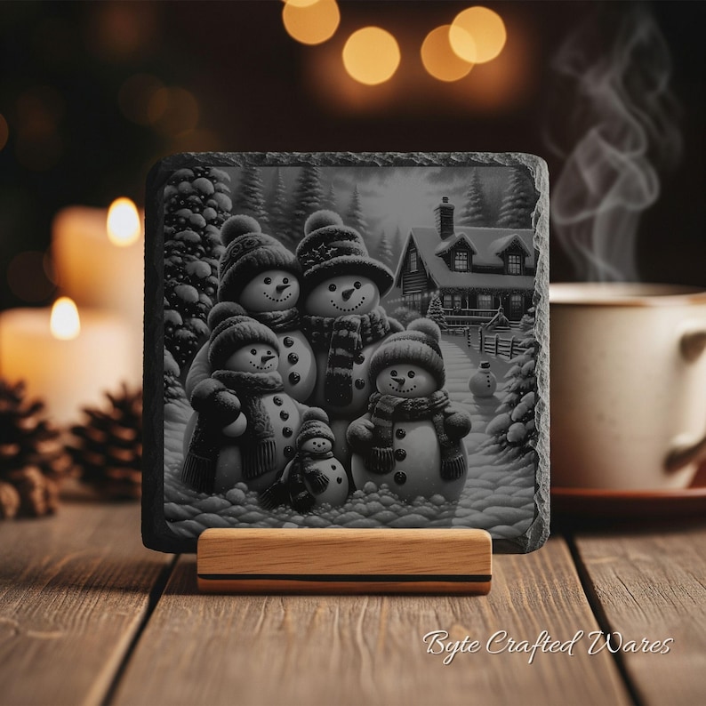 May include: A square slate coaster featuring a black and white illustration of a family of five snowmen wearing hats and scarves in a snowy landscape with a house in the background, resting on a light-colored wooden display stand.