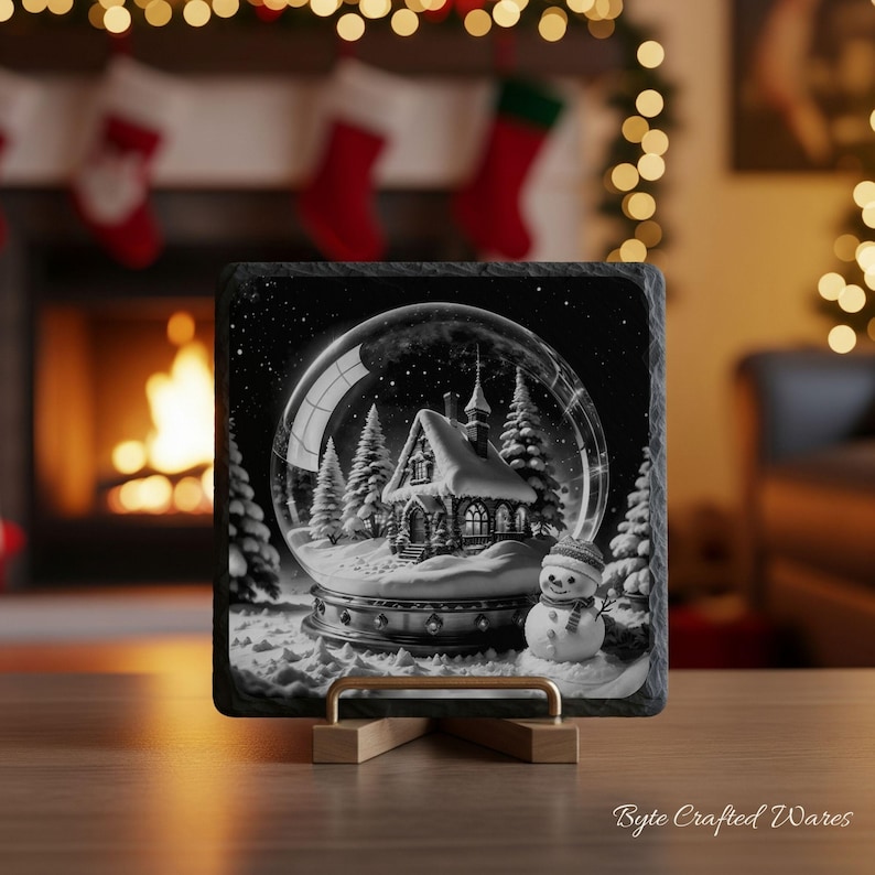 May include: A square slate coaster featuring a black and white illustration of a snow globe containing a house and pine trees, with a snowman standing in the foreground. The coaster is displayed on a small wooden stand with a textured edge.