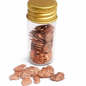 May include: A clear glass vial with a gold-colored cap filled with irregularly shaped, reddish-brown copper flakes. Several flakes are scattered around the base of the vial on a white background. The flakes have a textured appearance.